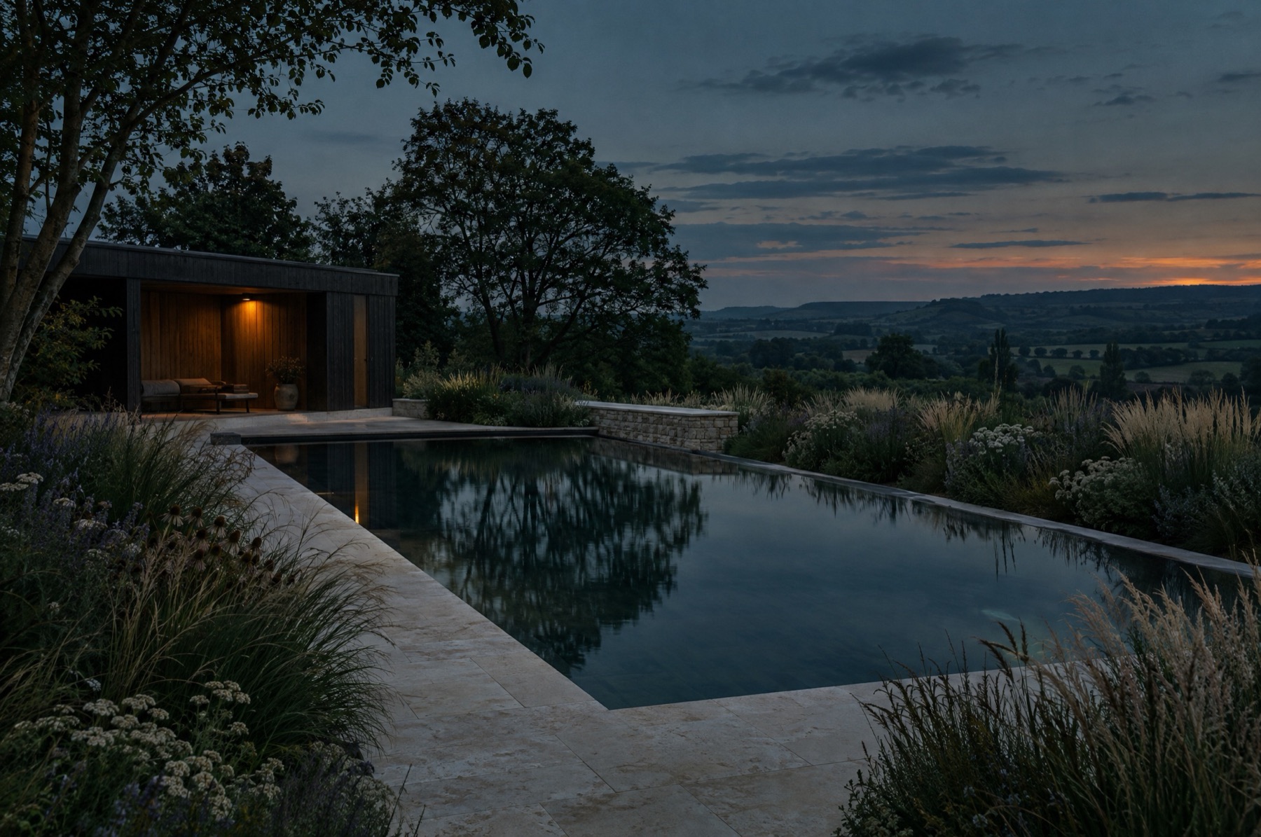 A natural swimming pool terrace at dusk, dark still water lined in stone with a lit timber pool house.